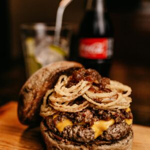 Close-up of a delicious gourmet burger topped with onion rings, cheese, and served with a bottle of cola.